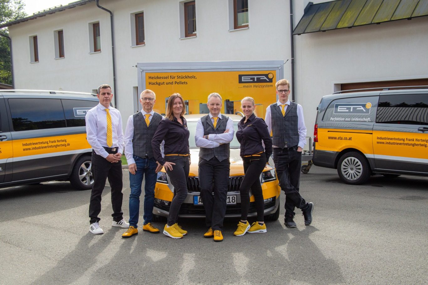 Gruppe von fünf Personen in gelb-schwarzer Kleidung vor Fahrzeugen, freundlich lächelnd.